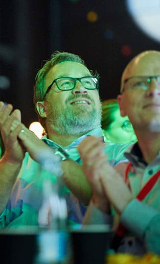Mand smiler og klapper under en konference i Roskilde Kongres- og Idrætscenter på Sjælland, hvor deltagerne nyder oplæg og fællesskab.