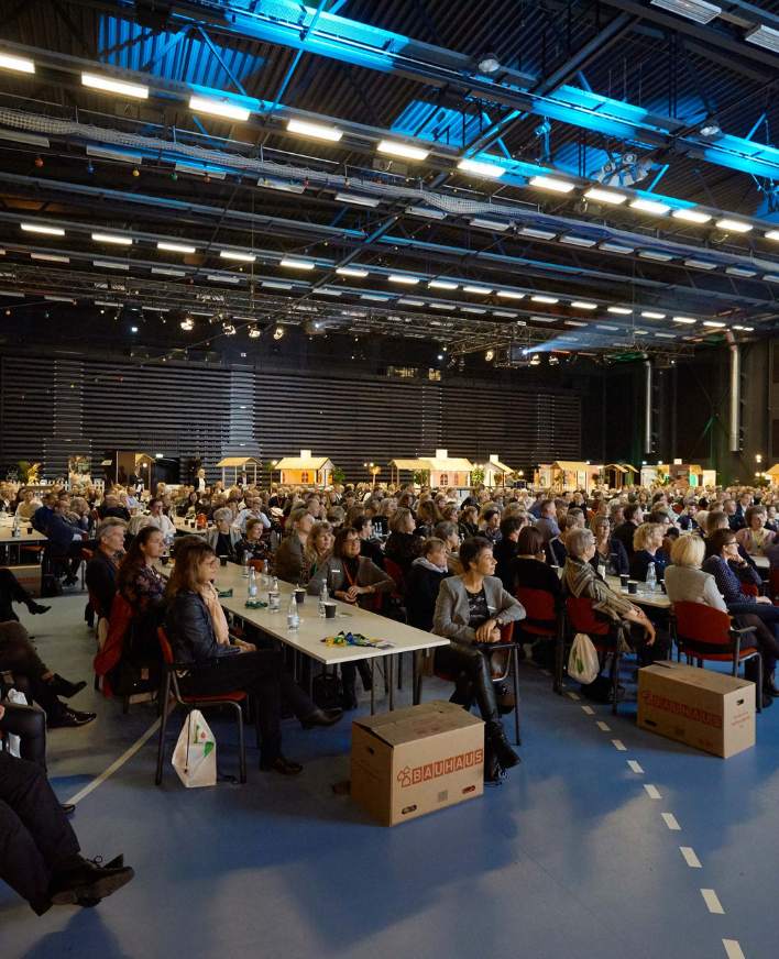 Firmaevent i stor hal med scene, skærme og deltagere ved bordopstilling